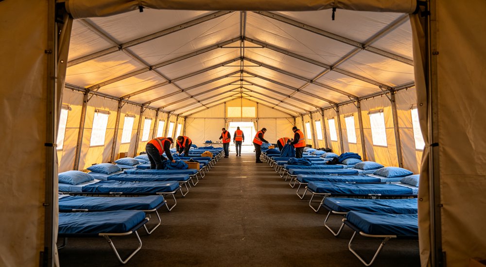 Eye-level shot of large emergency relief tent interior with rows of blue cots and white bedding carefully arranged for disaster response victims in temporary accommodation
